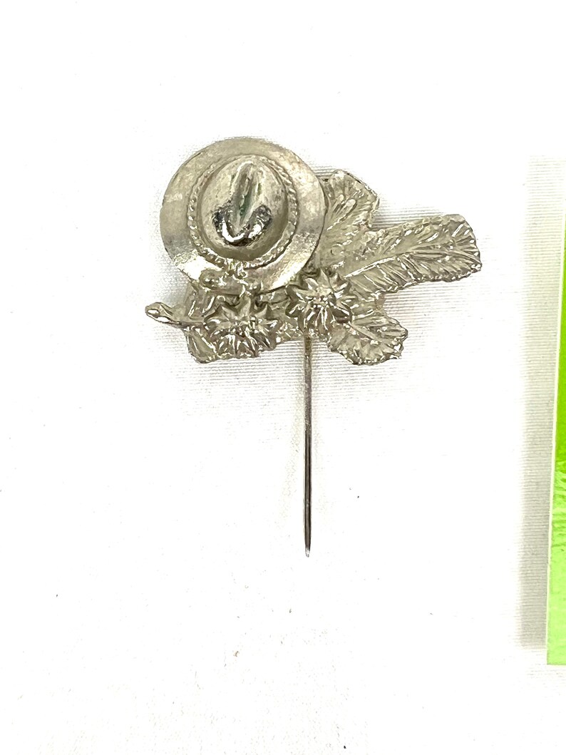 May include: Silver-toned pin featuring a stylized hat with a flower design. The pin is attached to a long, thin pin.