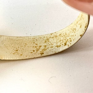 May include: Close-up of a curved, off-white metal object with a textured, speckled surface. The object has a slightly tarnished appearance with areas of brown discoloration. The background is a plain white surface.