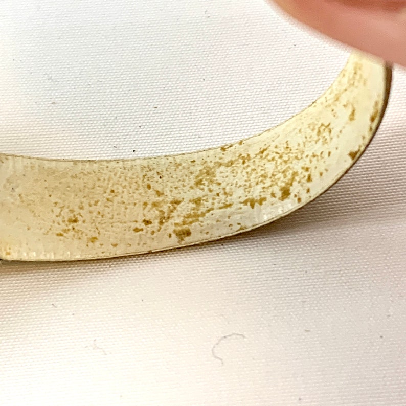 May include: Close-up of a curved, off-white metal object with a textured, speckled surface. The object has a slightly tarnished appearance with areas of brown discoloration. The background is a plain white surface.