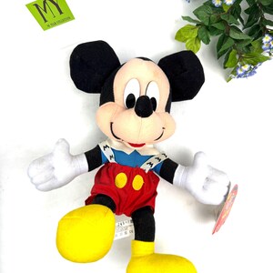 May include: A plush Mickey Mouse toy with black ears, a tan face, and white gloves. He wears a blue shirt, red shorts, and yellow shoes. The toy has a tag attached and is set against a white background. The toy is from the "MY" collection.