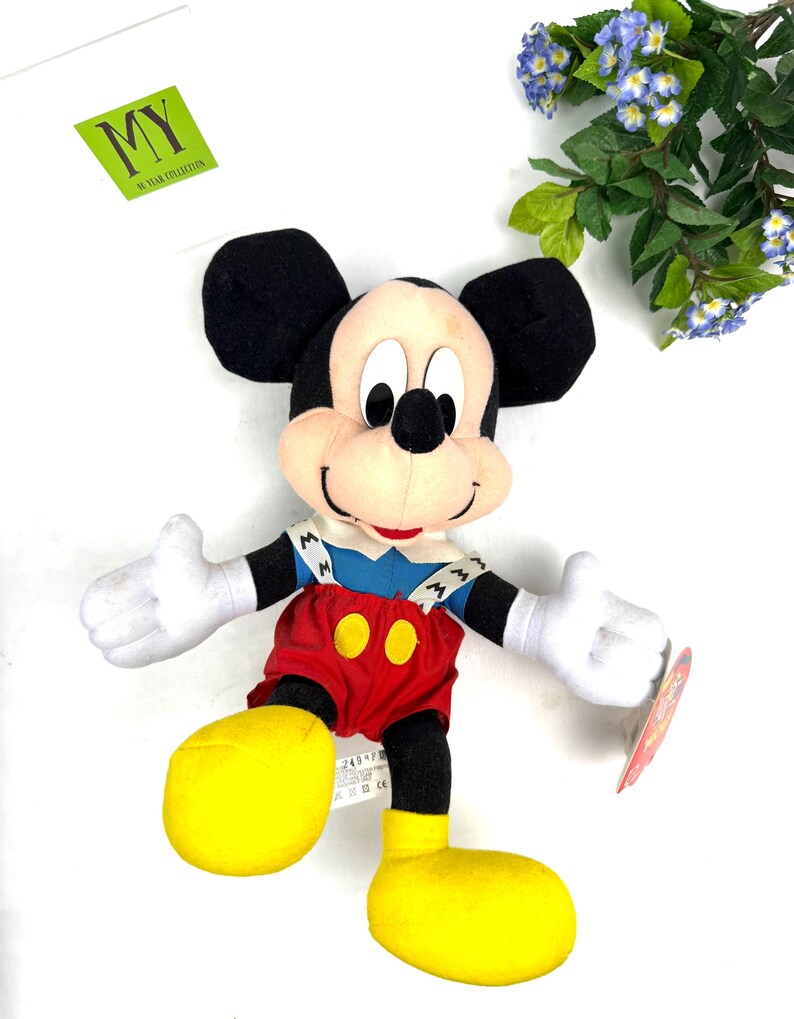 May include: A plush Mickey Mouse toy with black ears, a tan face, and white gloves. He wears a blue shirt, red shorts, and yellow shoes. The toy has a tag attached and is set against a white background. The toy is from the "MY" collection.