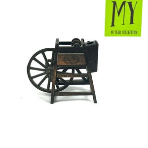 May include: A vintage Play-Me pencil sharpener in the shape of a wooden wheel with a metal crank. The sharpener is brown and black and has a metal blade. The sharpener is on a white background.