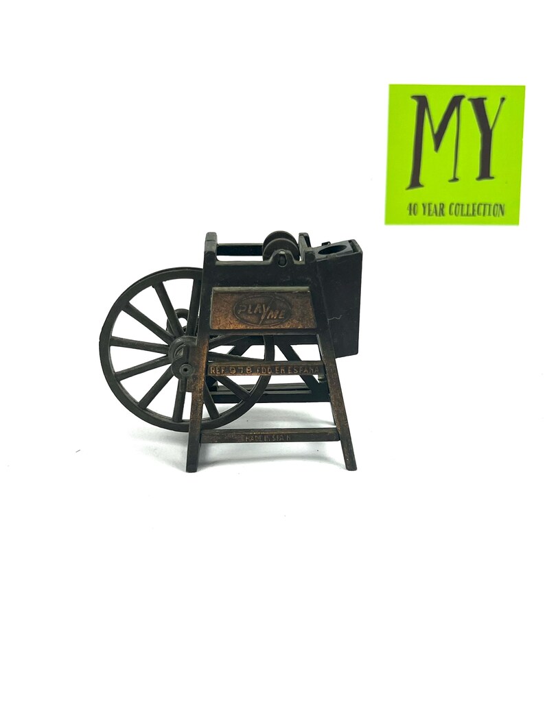 May include: A vintage Play-Me pencil sharpener in the shape of a wooden wheel with a metal crank. The sharpener is brown and black and has a metal blade. The sharpener is on a white background.