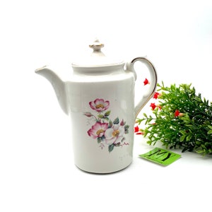 Può includere: Teiera in ceramica bianca con un design floreale con fiori rosa e bianchi. La teiera ha un bordo dorato attorno al coperchio e al manico.