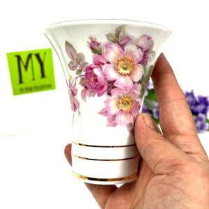May include: A white ceramic vase with a flared opening and a floral design. The vase features pink and white roses with green leaves. The rim of the vase has a gold trim.