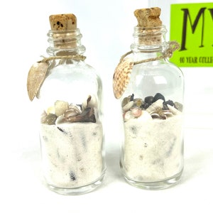 May include: Two small glass bottles filled with white sand and seashells. The bottles have cork stoppers and are tied with twine. The bottles are on a white surface.