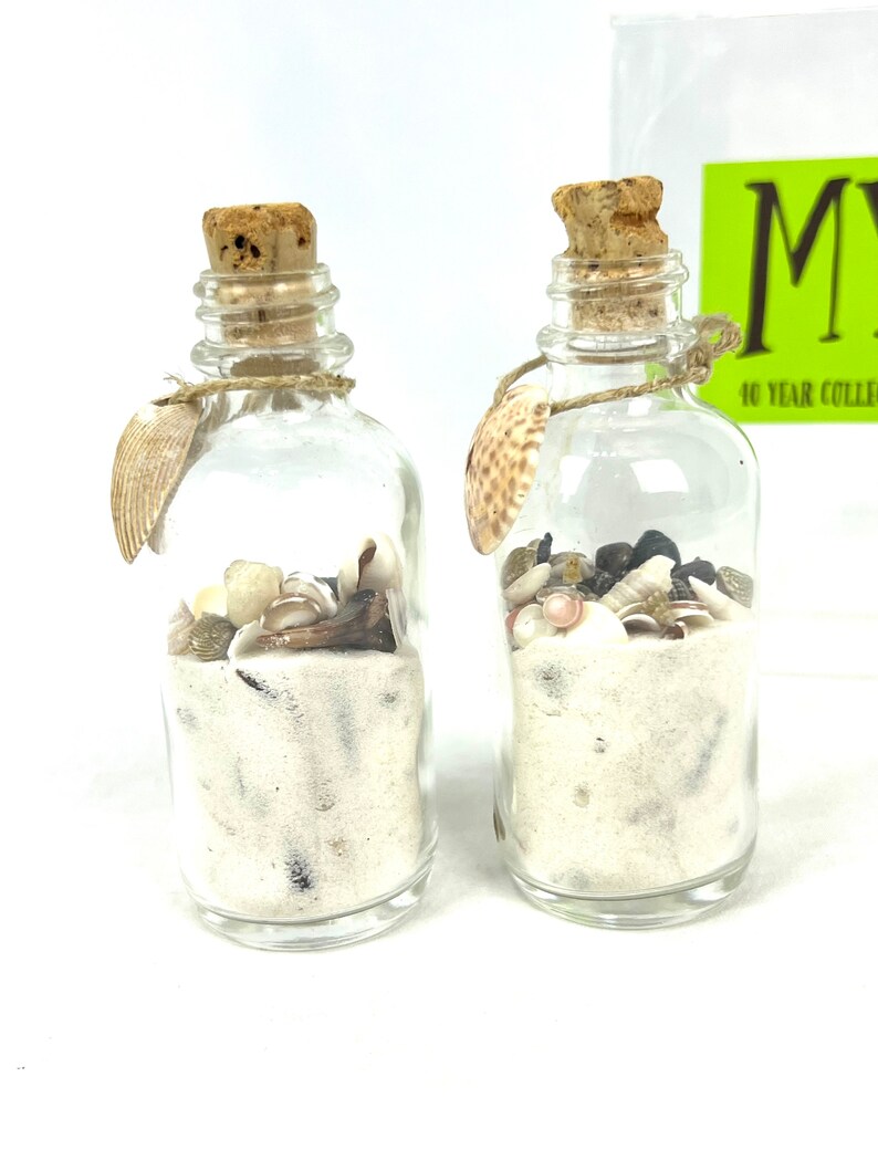 May include: Two small glass bottles filled with white sand and seashells. The bottles have cork stoppers and are tied with twine. The bottles are on a white surface.