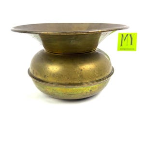 May include: A vintage brass bowl with a wide, shallow top and a rounded base. The bowl has a tarnished, aged appearance, with a greenish hue in some areas. The text "MY" is visible on a green square.