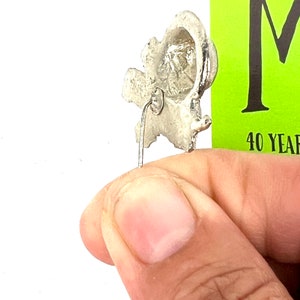 May include: Silver-toned metal pin with a detailed design of a stylized ear. The pin has a straight back pin with a loop at the top.