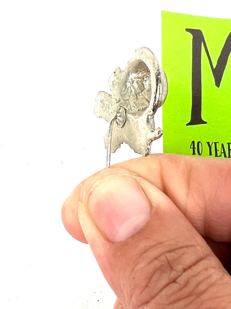 May include: Silver-toned metal pin with a detailed design of a stylized ear. The pin has a straight back pin with a loop at the top.