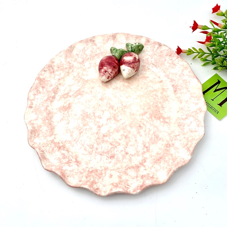 May include: A pink ceramic plate with a scalloped edge and a white marbled design. Two small red and white ceramic radishes with green leaves are on the plate.