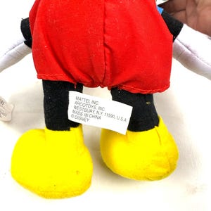 May include: A Mickey Mouse plush toy, viewed from the back, featuring a red shorts, black legs, and yellow shoes. The toy has white gloves and a black head. A tag with text is attached to the toy.