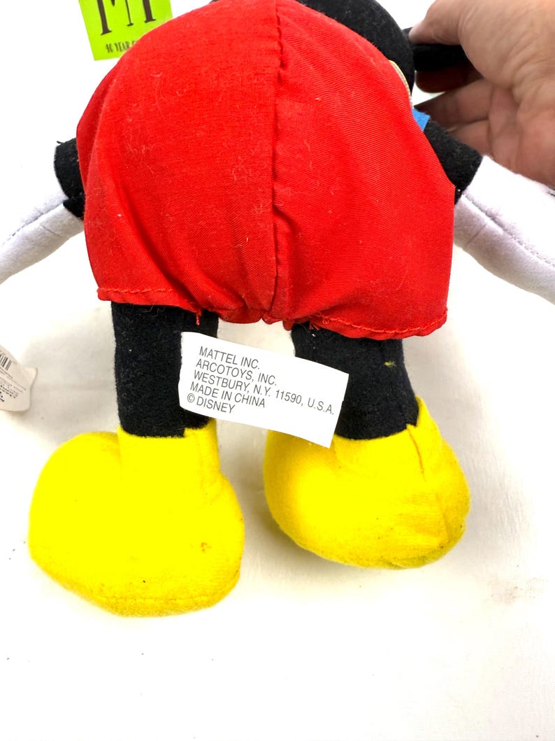 May include: A Mickey Mouse plush toy, viewed from the back, featuring a red shorts, black legs, and yellow shoes. The toy has white gloves and a black head. A tag with text is attached to the toy.