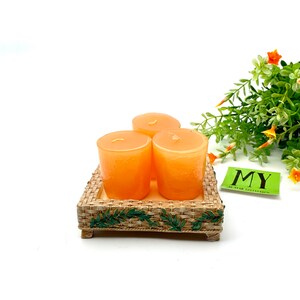 May include: Three orange votive candles on a woven tray with a green leaf design. The tray has a rope trim and is sitting on a white surface.