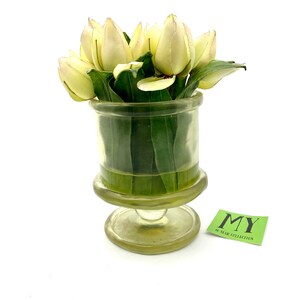 May include: A clear glass vase with a pedestal base holds a bouquet of white tulips with green leaves. The vase has a textured surface and the tulips are arranged in a cluster.