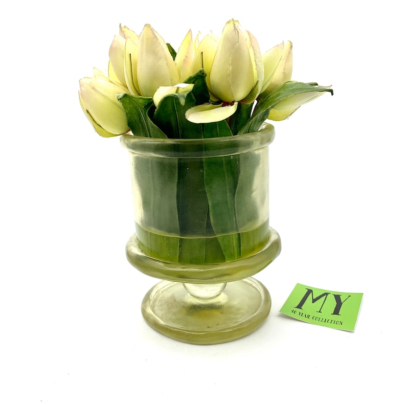 May include: A clear glass vase with a pedestal base holds a bouquet of white tulips with green leaves. The vase has a textured surface and the tulips are arranged in a cluster.