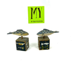 May include: Two wooden fish figurines with painted details, each standing on a small wooden cube with painted fish designs. The cubes are painted black with white, yellow, and green fish. The fish figurines are painted gray with white spots and have black eyes. The text "MY 40 YEAR COLLECTION" is on a yellow sticker.