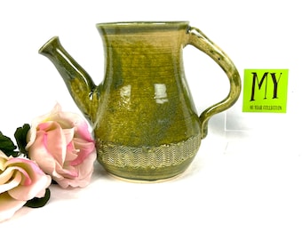 May include: A green ceramic teapot with a textured base and a handle. The teapot has a spout and a slightly rounded body.