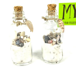 May include: Two small glass bottles filled with sand, seashells, and small stones. The bottles have cork stoppers and are tied with twine. The bottles are on a white surface.