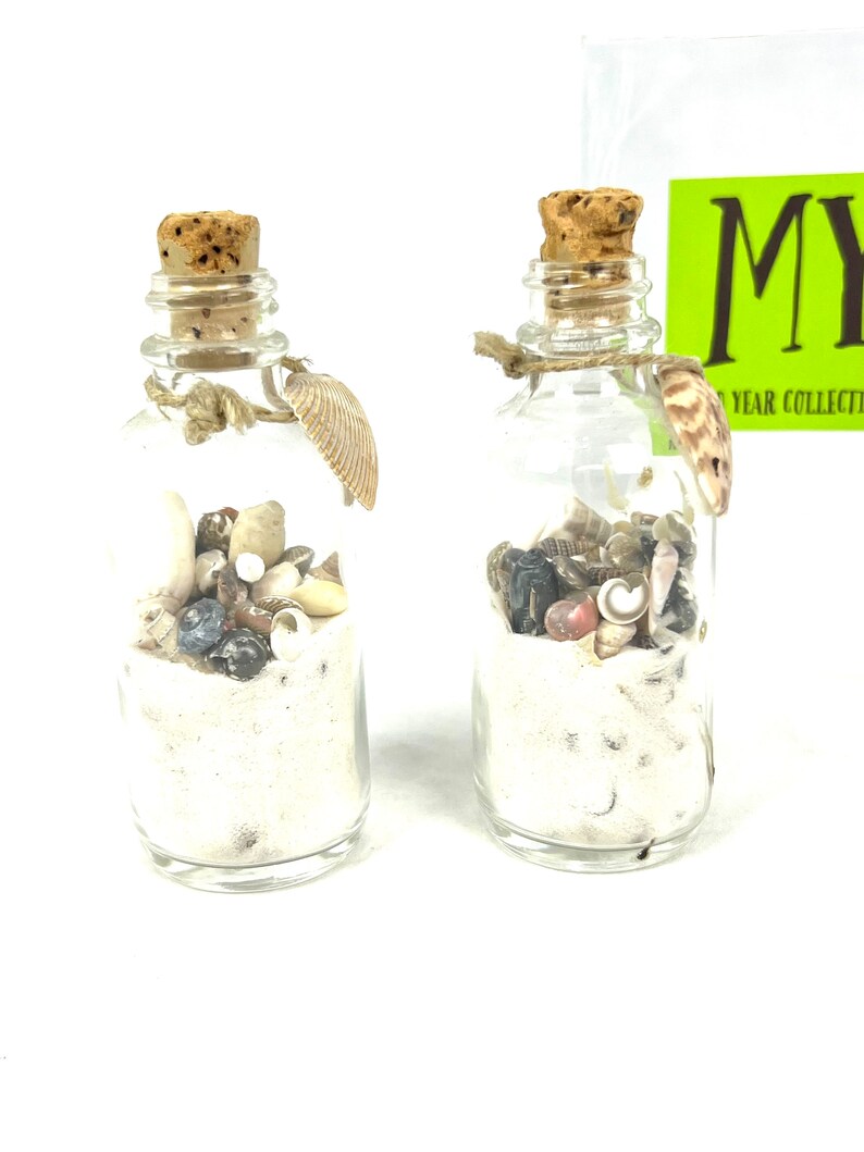 May include: Two small glass bottles filled with sand, seashells, and small stones. The bottles have cork stoppers and are tied with twine. The bottles are on a white surface.
