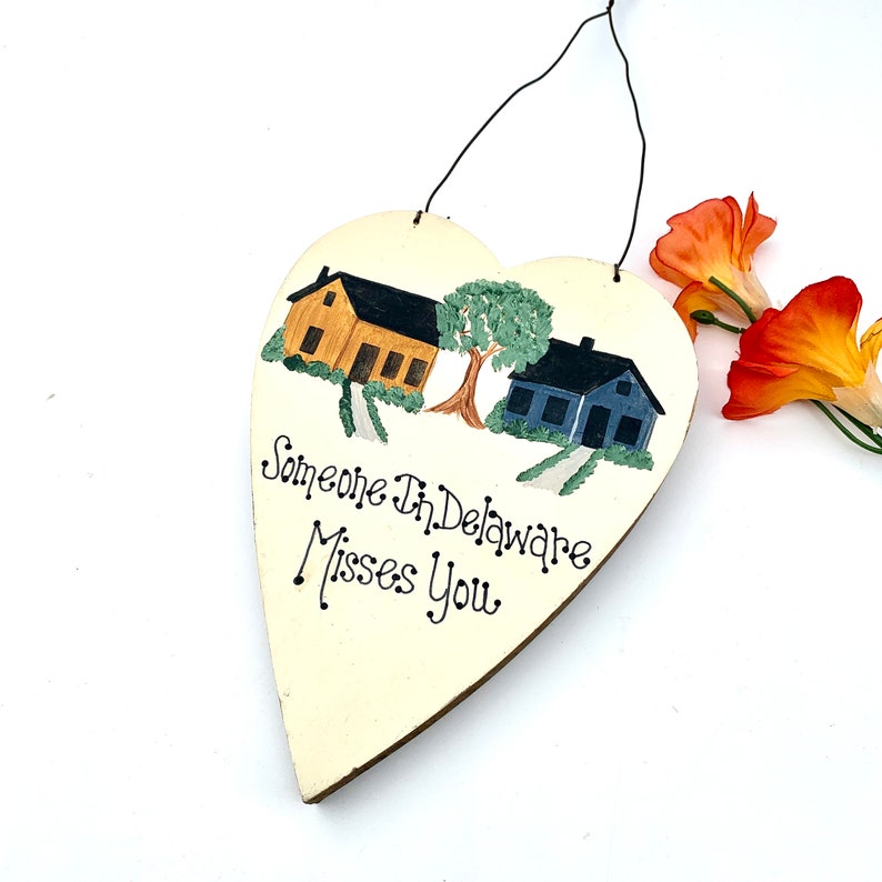 May include: A white heart-shaped wooden sign with a painted scene of two houses and a tree. The text "Someone In Delaware Misses You" is written in black cursive.
