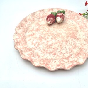 May include: A pink and white ceramic serving plate with a scalloped edge. The plate has a marbled design and features two small, white ceramic radishes with green tops.