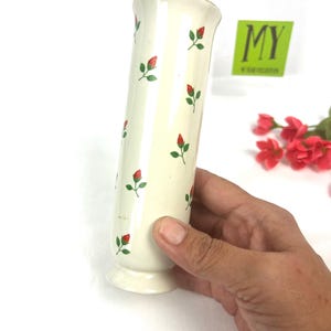 May include: White ceramic vase with a red and green floral pattern. The vase is cylindrical and has a flared lip.