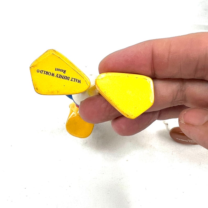 Może przedstawiać: Ż&oacute;łta plastikowa figurka Donalda Kaczora w niebiesko-białym stroju marynarskim. Figurka stoi na nogach i ma napis "WALT DISNEY WORLD&reg; Resort" na spodzie.