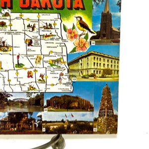May include: A vintage postcard featuring a map of North Dakota with various landmarks and points of interest. The map is surrounded by smaller images of different locations in the state, including a church, a building, and a monument.