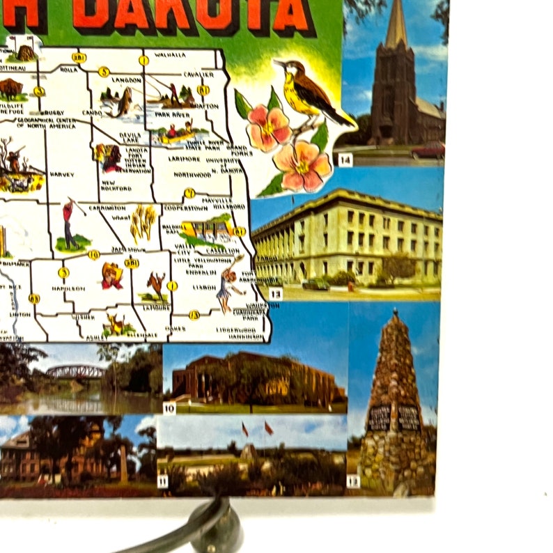 May include: A vintage postcard featuring a map of North Dakota with various landmarks and points of interest. The map is surrounded by smaller images of different locations in the state, including a church, a building, and a monument.