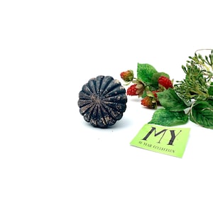 Könnte beinhalten: Ein schwarzer Metallknopf mit einem dekorativen, sonnenstrahlenartigen Design. Der Knopf hat eine abgenutzte Oberfläche, die ihm einen Vintage-Look verleiht.