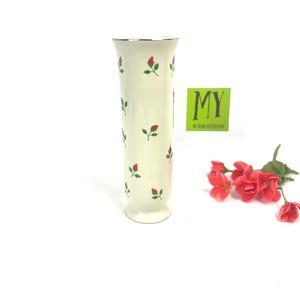 May include: A white ceramic vase with a gold rim and a pattern of red and green roses. The vase has a flared base and a cylindrical body.