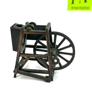 May include: A vintage metal pencil sharpener shaped like a spinning wheel. The sharpener is brown and black with a wooden design. It has a small black compartment for shavings and a metal wheel with spokes.