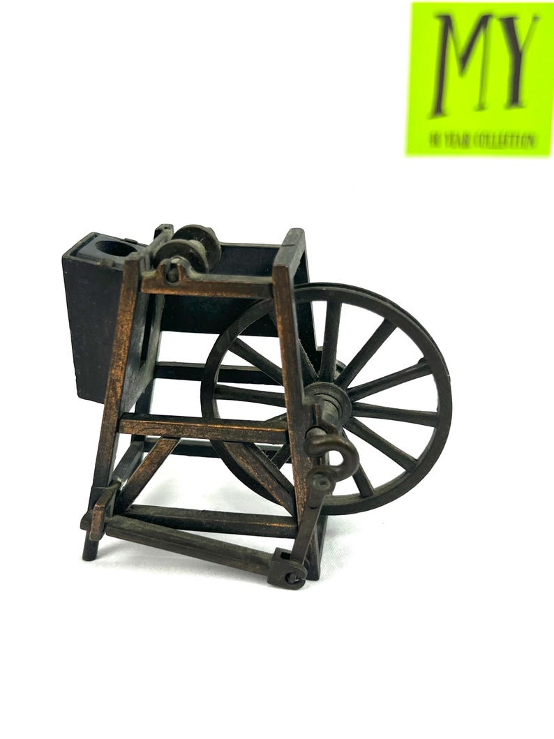 May include: A vintage metal pencil sharpener shaped like a spinning wheel. The sharpener is brown and black with a wooden design. It has a small black compartment for shavings and a metal wheel with spokes.