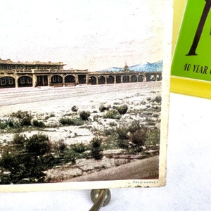 May include: Vintage postcard depicting a landscape with a long, arched building and a mountain range in the background. The postcard has a sepia tone with touches of colour. The bottom right corner has the text "FRED HARVEY".