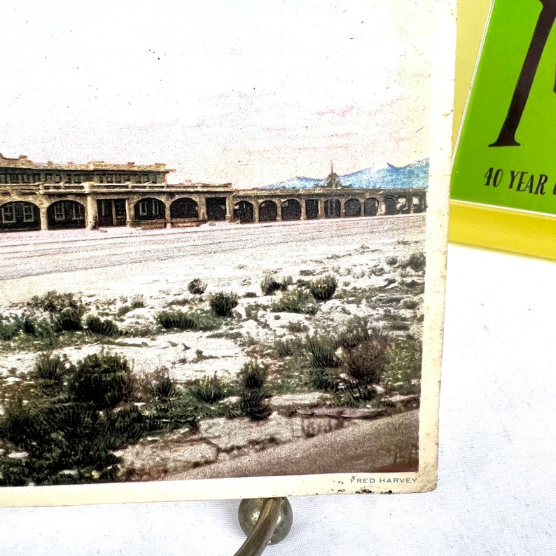 May include: Vintage postcard depicting a landscape with a long, arched building and a mountain range in the background. The postcard has a sepia tone with touches of colour. The bottom right corner has the text "FRED HARVEY".