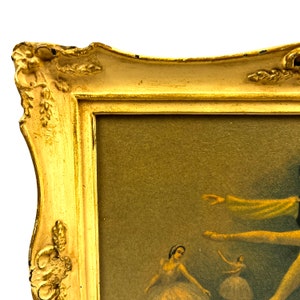 May include: A gold-colored ornate frame with a picture of ballerinas in a vintage style. The frame has a raised, decorative edge and a light brown mat.
