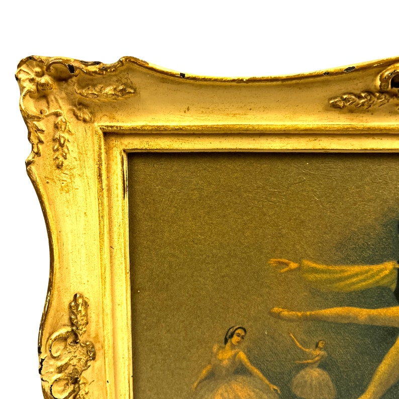 May include: A gold-colored ornate frame with a picture of ballerinas in a vintage style. The frame has a raised, decorative edge and a light brown mat.