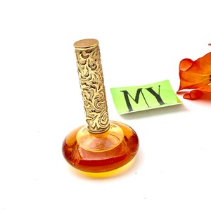 May include: A vintage amber glass perfume bottle with a gold, ornate stopper. The bottle has a round, flat base and a slender neck.