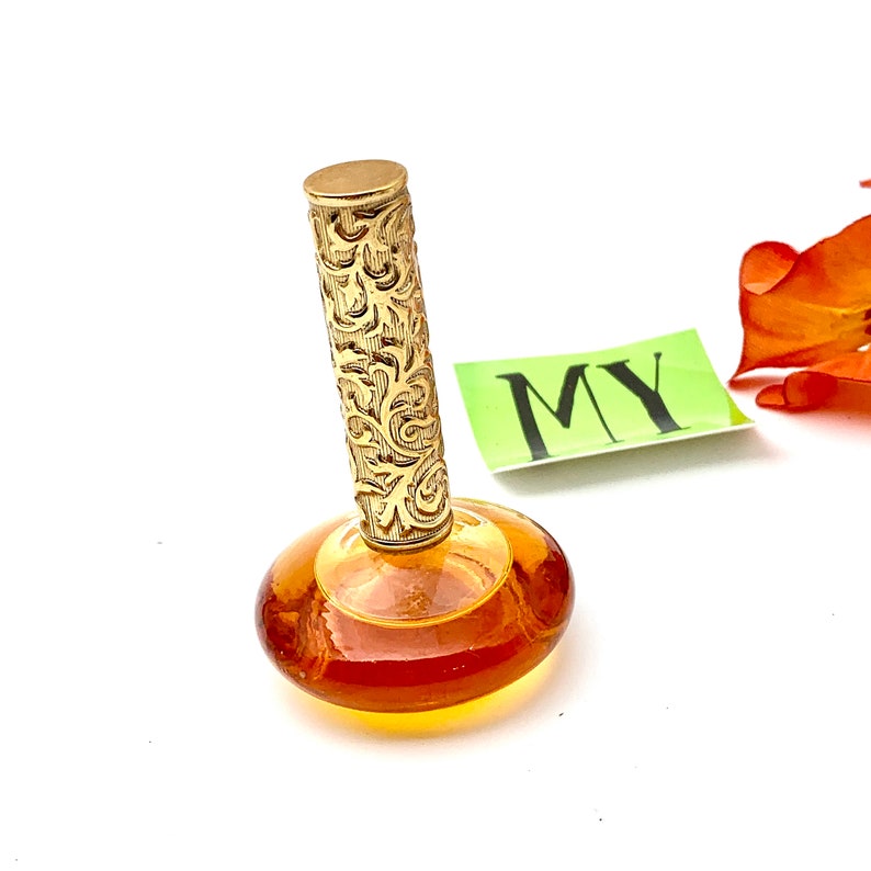 May include: A vintage amber glass perfume bottle with a gold, ornate stopper. The bottle has a round, flat base and a slender neck.