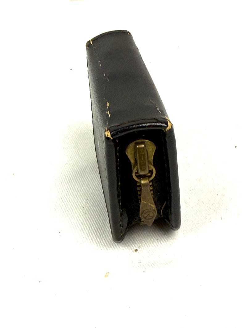 May include: A black leather wallet with a brass zipper. The rectangular wallet is closed, with the zipper pull at the bottom. The leather shows some wear and tear, with slight discoloration and scuff marks. The wallet is set against a white background.
