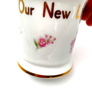 以下が含まれることがあります： 金色の縁取りと「Our New」という金色の文字が書かれた白いセラミック製のマグカップ。マグカップにはピンクの花の模様が描かれています。