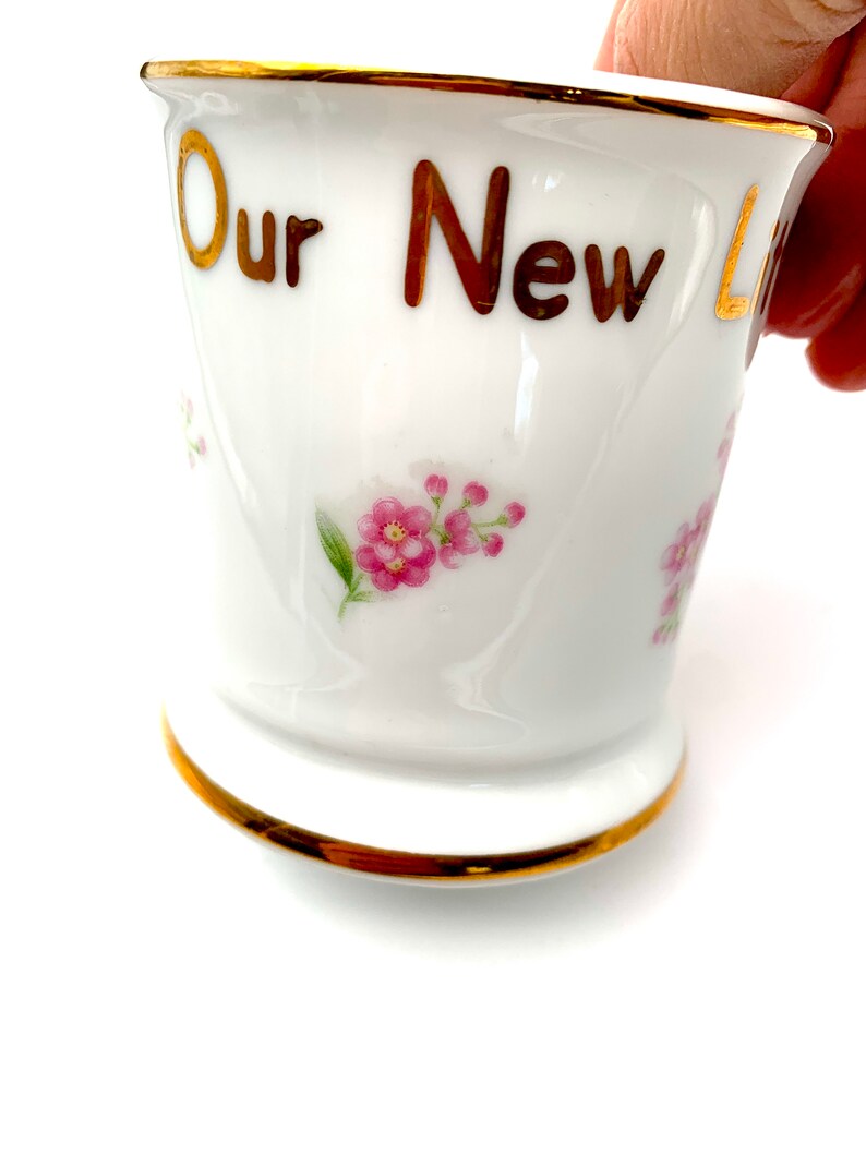 以下が含まれることがあります： 金色の縁取りと「Our New」という金色の文字が書かれた白いセラミック製のマグカップ。マグカップにはピンクの花の模様が描かれています。