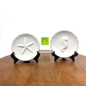 May include: Two white ceramic decorative plates with raised designs of a starfish and a seahorse. The plates are on small black stands and are displayed on a wooden surface.
