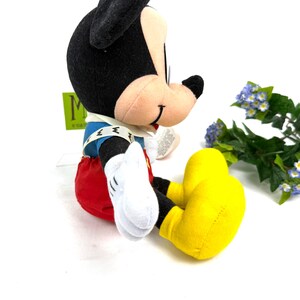 May include: A plush Mickey Mouse toy in a seated position. Mickey has black ears, a tan face, and wears a blue jacket, red shorts, yellow shoes, and white gloves. The toy is on a white surface with a green tag in the background.