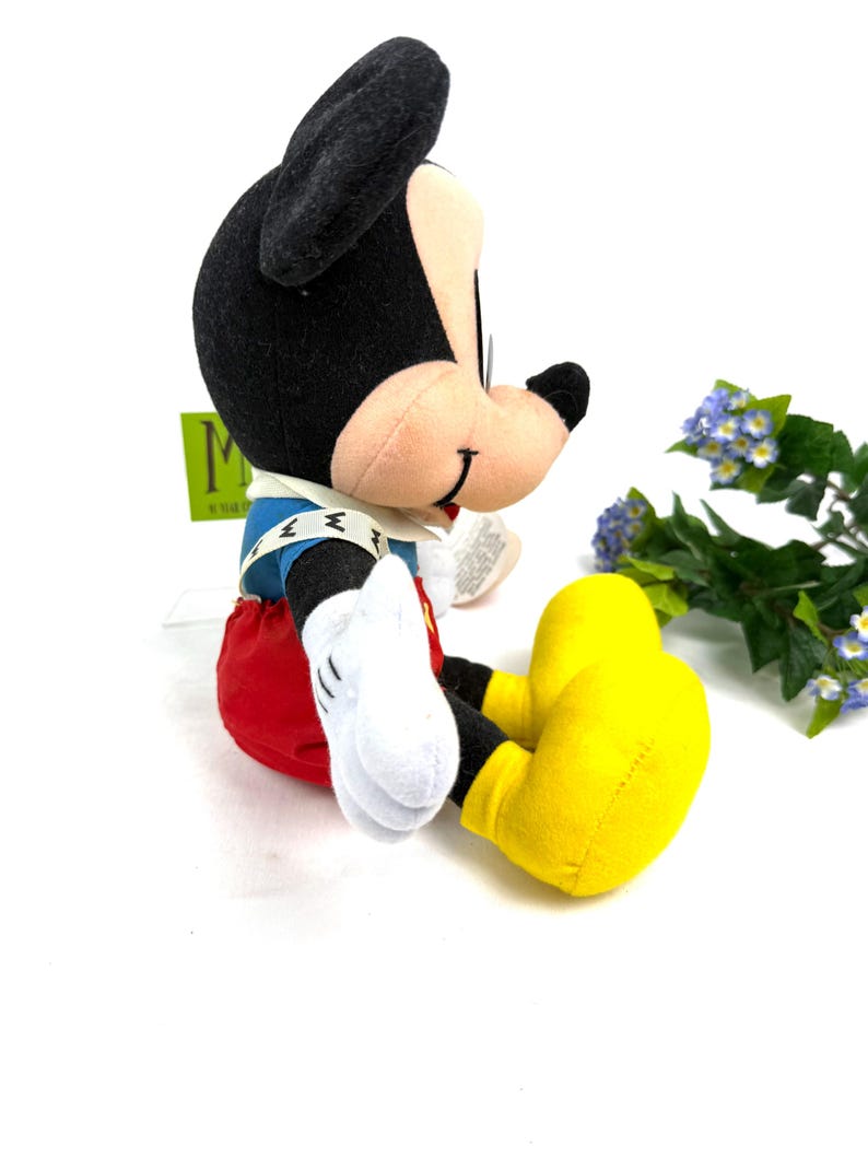 May include: A plush Mickey Mouse toy in a seated position. Mickey has black ears, a tan face, and wears a blue jacket, red shorts, yellow shoes, and white gloves. The toy is on a white surface with a green tag in the background.