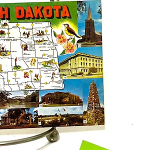 May include: A vintage postcard featuring a map of North Dakota with various landmarks and points of interest. The postcard includes a map of the state, a church, a building, a monument, and a scenic view. The postcard is in good condition with some wear and tear.