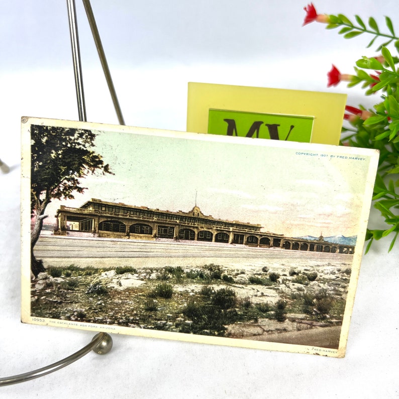 May include: Aged postcard depicting a long building with arched openings, possibly a railway station. The image has a faded colour scheme, with a desert scene in the foreground. Text reads "The Escalante, Ash Fork, Arizona."