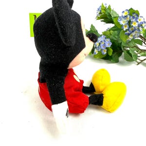 May include: A plush Mickey Mouse toy, featuring a black head and ears, red shorts, white gloves, and yellow shoes. The toy is sitting down, with its arms and legs extended. The background is white.