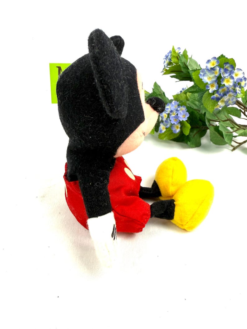 May include: A plush Mickey Mouse toy, featuring a black head and ears, red shorts, white gloves, and yellow shoes. The toy is sitting down, with its arms and legs extended. The background is white.